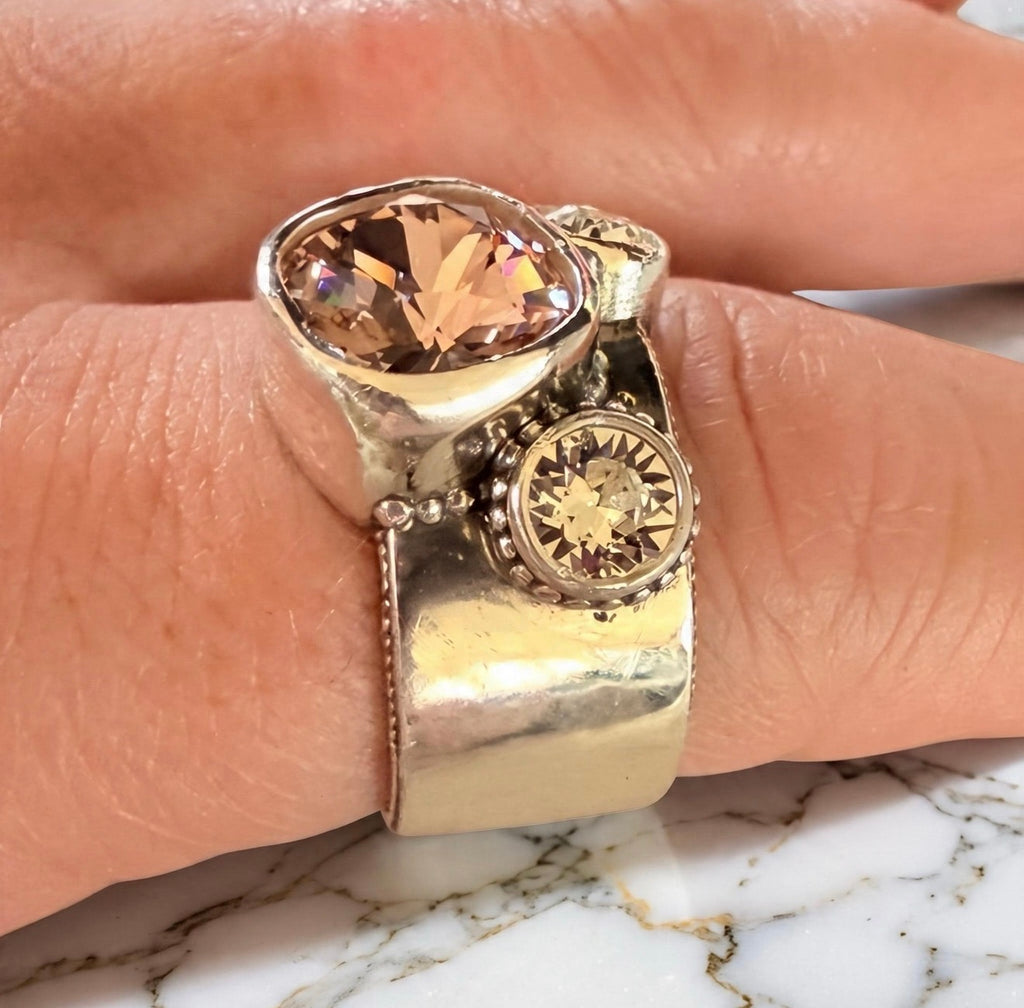 A close-up of a hand wearing a wide, chunky silver ring with a large peach-pink colored cushion cut Swarovski crystal and a smaller round crystal accent.