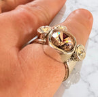 A close-up of a hand wearing a chunky silver ring with a large faceted iridescent peach/pink Swarovski crystal and smaller accent stones.