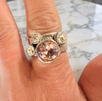A silver ring with a large cushion-cut peach-pink center stone and three smaller round clear accent stones, shown worn on a finger against a marble surface.