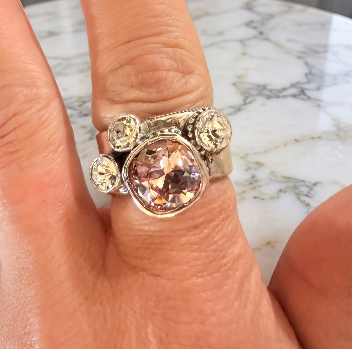 A silver ring with a large cushion-cut peach-pink center stone and three smaller round clear accent stones, shown worn on a finger against a marble surface.